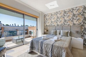 A bedroom with a large bed, a nightstand, a lamp, a window with a view of buildings, and a balcony with a table and chairs.