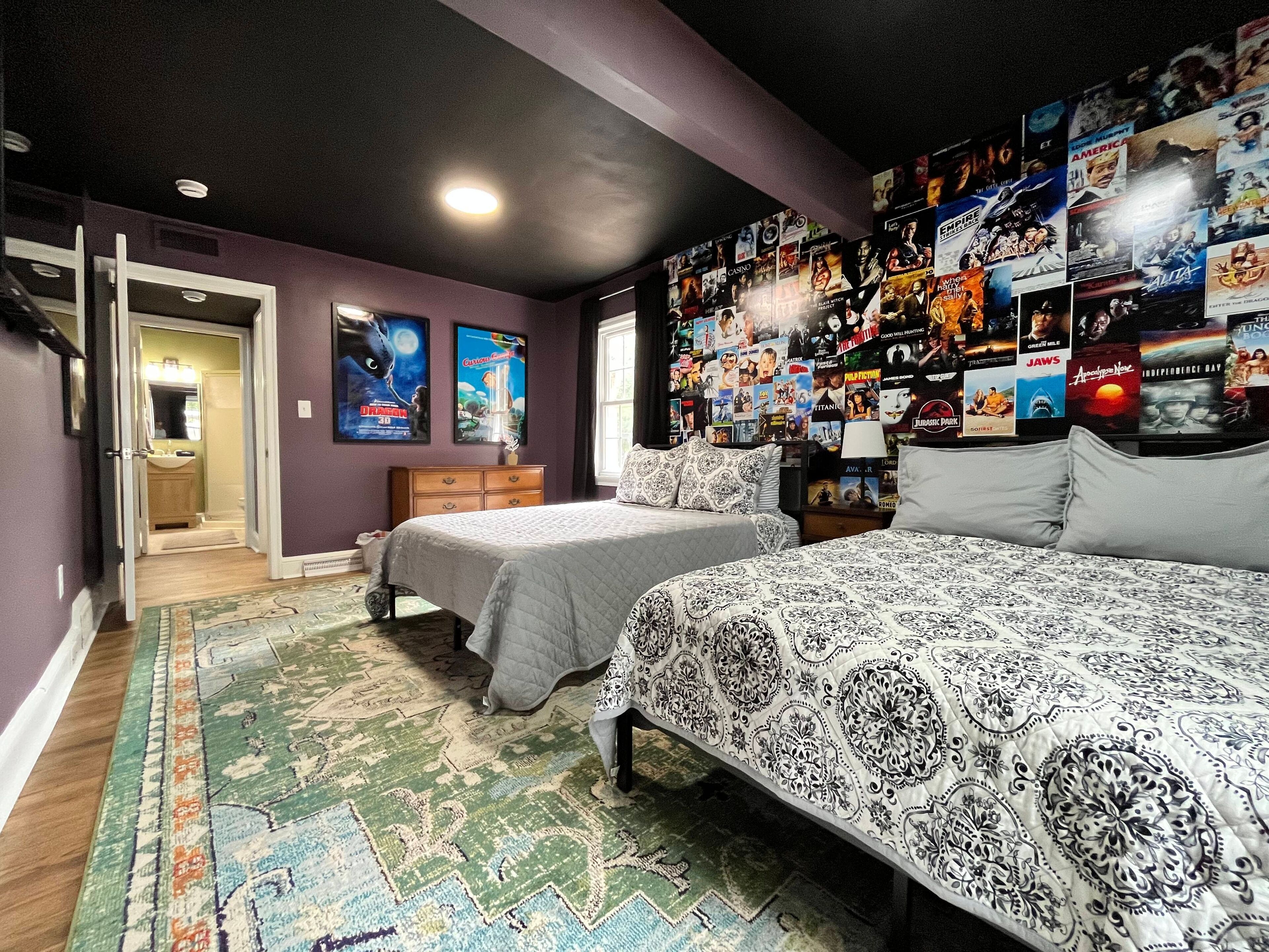 Un dormitorio con dos camas, una pared llena de carteles de películas y un baño visible a través de una puerta abierta.