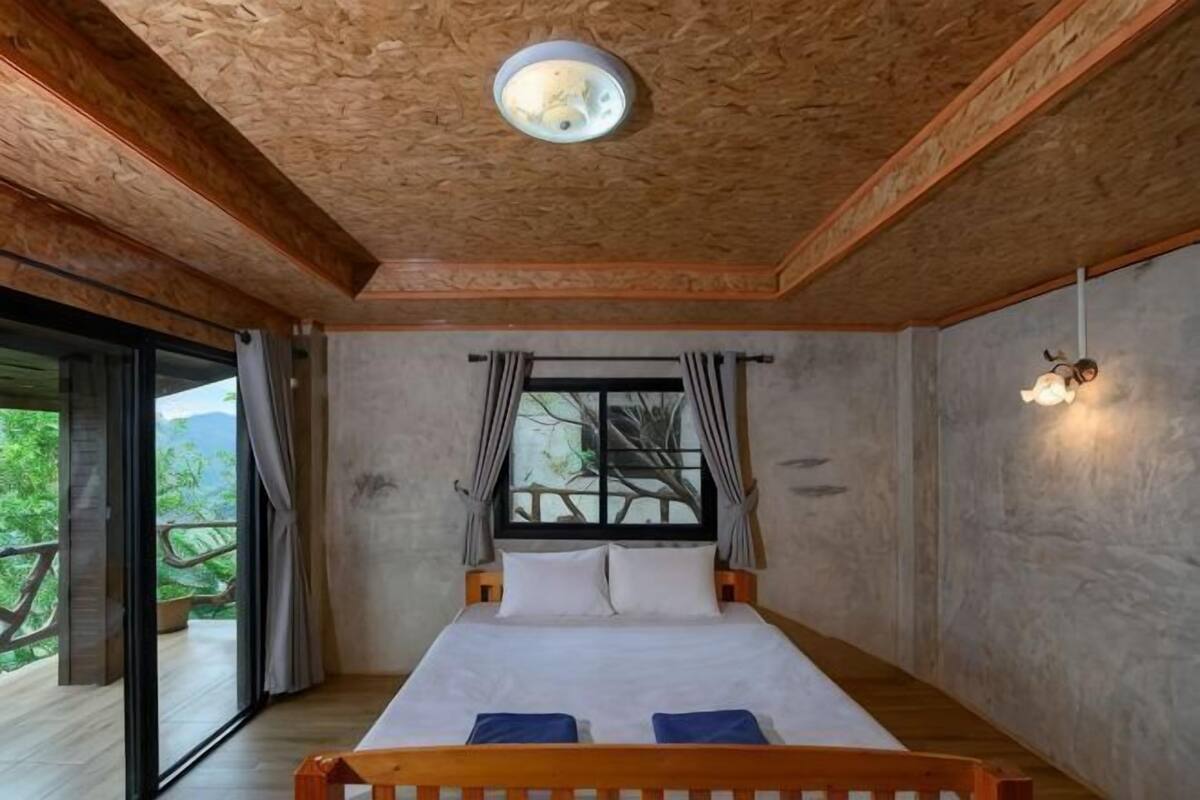 A bedroom with a wooden bed, a window with curtains, and a view of greenery outside.