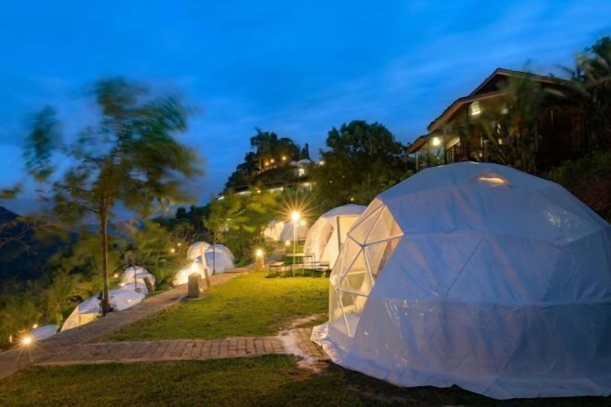 A series of dome-shaped tents with a pathway leading to a building, surrounded by trees and a mountainous backdrop.