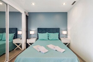 Un lit bien fait, recouvert d’une couverture verte, deux tables de chevet avec des lampes et un grand miroir sur un des murs.
