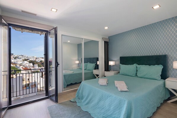 A bedroom with a balcony, a bed with a green bedspread, a nightstand, a bedside table, and a mirror.