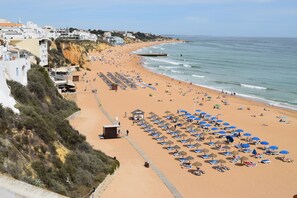 A beach with rows of sunbeds and umbrellas, a lifeguard tower, and buildings along the coastline.