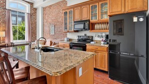 A kitchen with granite countertops, wooden cabinets, a black refrigerator, and a brick wall. - Savannah Serendipity | Historic Broughton St (Savannah)