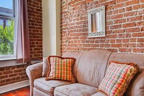 A room with a brick wall, a window with curtains, a sofa with plaid cushions, and a mirror.