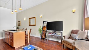 A living room with a kitchen area, wooden flooring, a flat-screen TV, and a dining table with chairs. - Savannah Serendipity | Historic Broughton St (Savannah)