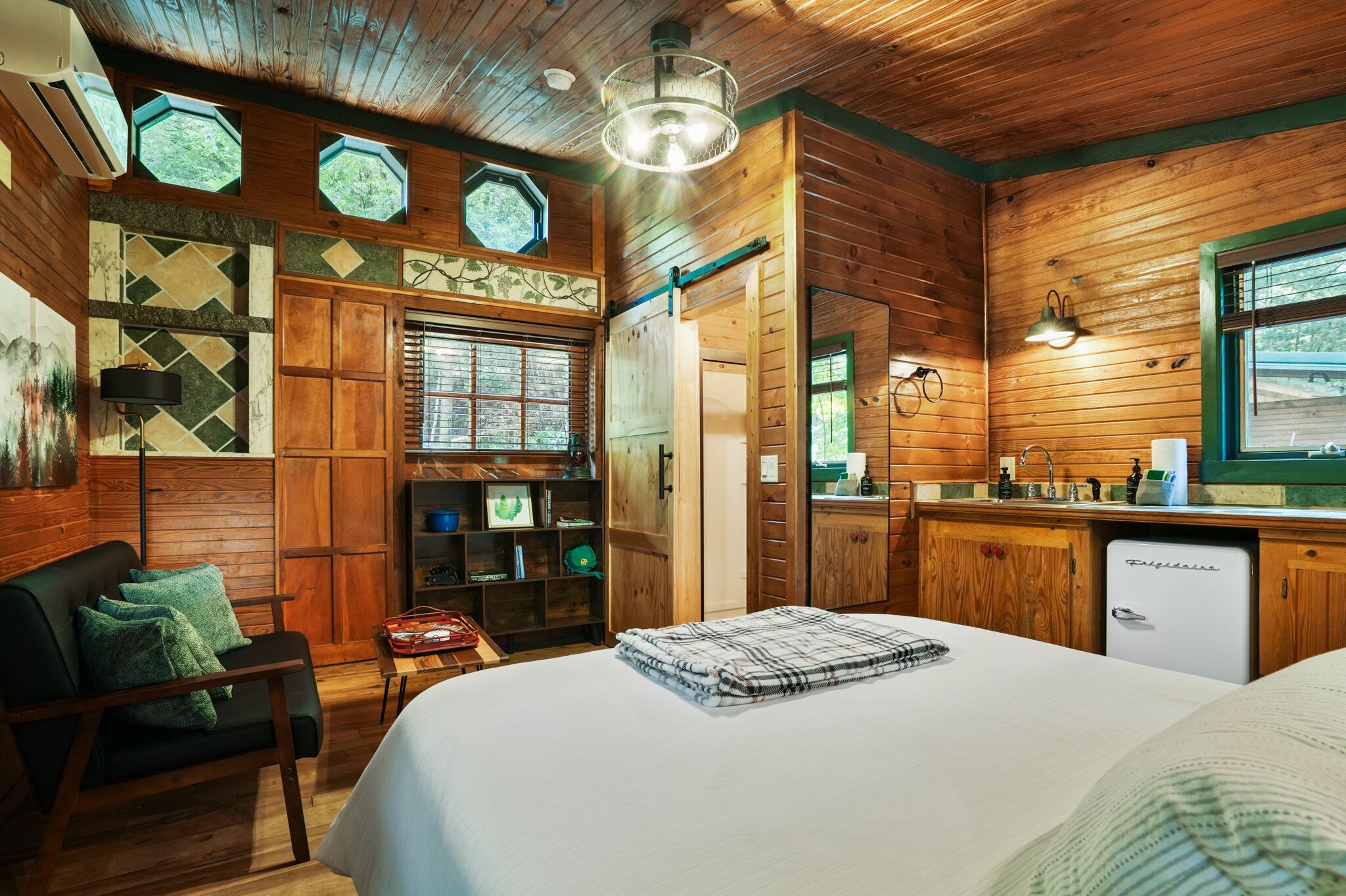 A cozy cabin-style room with a bed, a desk, a chair, and a kitchenette.