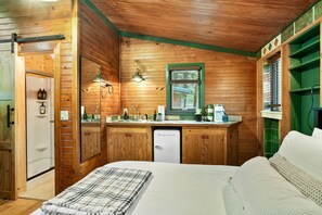 A wooden cabin-style room with a bed, a small kitchenette, and a window with a view of greenery.