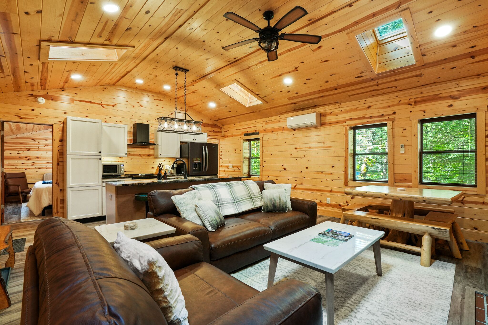 Interior acogedor de una cabaña con sala de estar, cocina y comedor, con paredes de madera, ventilador de techo y ventanas grandes.