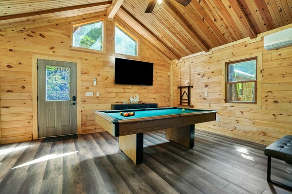 A wooden cabin with a pool table, a television mounted on the wall, and large windows.