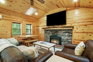 A cozy cabin interior with a stone fireplace, wooden walls, a leather sofa, and a flat-screen TV mounted above the fireplace.