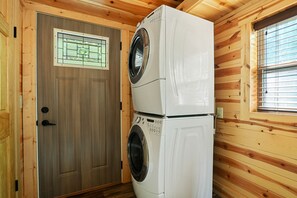 Ein Zimmer mit Holzwänden und einer Tür mit Fenster. Zwei übereinander gestellte Geräte sind zu sehen, eines scheint eine Waschmaschine und das andere ein Trockner zu sein.