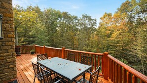 A wooden deck with a table and chairs, surrounded by a stone wall and a wooden railing, overlooking a forested area.