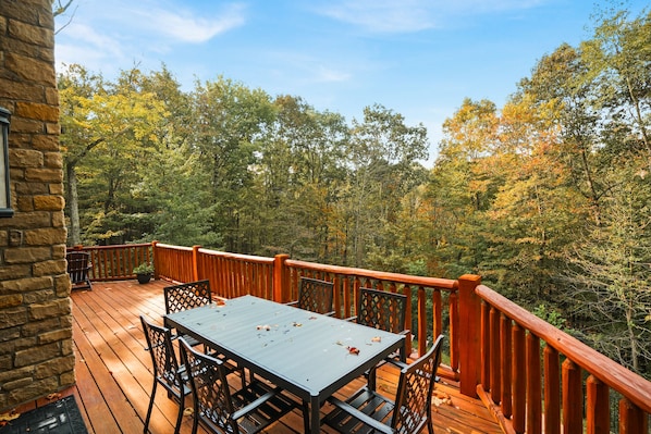 A wooden deck with a table and chairs, surrounded by a stone wall and a wooden railing, overlooking a forested area.