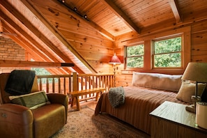 A cozy cabin with a loft bedroom, wooden beams, a fireplace, a bench, and a window with a view of trees.