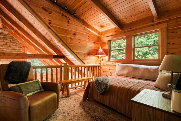 A cozy cabin with a loft bedroom, wooden beams, a fireplace, a bench, and a window with a view of trees.