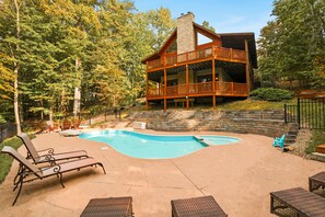 A two-story wooden cabin with a swimming pool, surrounded by trees.
