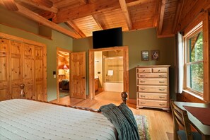 A bedroom with wooden furniture, a television, a bathroom with a shower, and a view through a window.