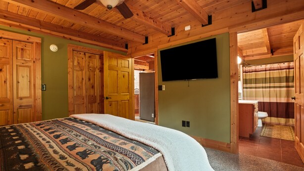 A wooden cabin bedroom with a bed, a television, and a bathroom visible through an open door.
