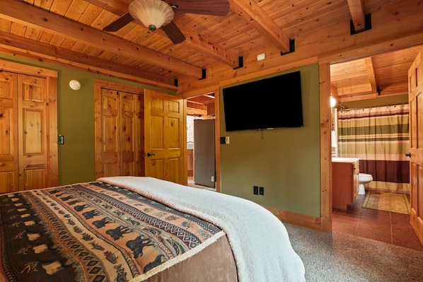 A wooden cabin bedroom with a bed, a television, and a bathroom visible through an open door.