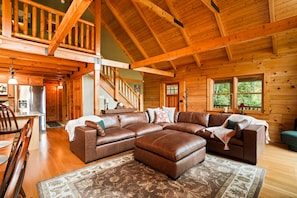A spacious log cabin interior with a large L-shaped sectional sofa, a dining area, and a staircase leading to an upper level.
