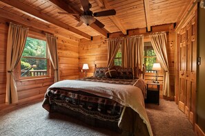 A log cabin bedroom with a bed, two windows with curtains, a ceiling fan, and a nightstand with a lamp. - Oasis Glen - Views, Pool, Hot Tub (Rockbridge)