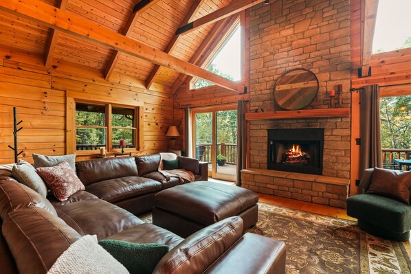 A log cabin interior with a large leather sectional sofa, a fireplace, and a view of the outdoors.