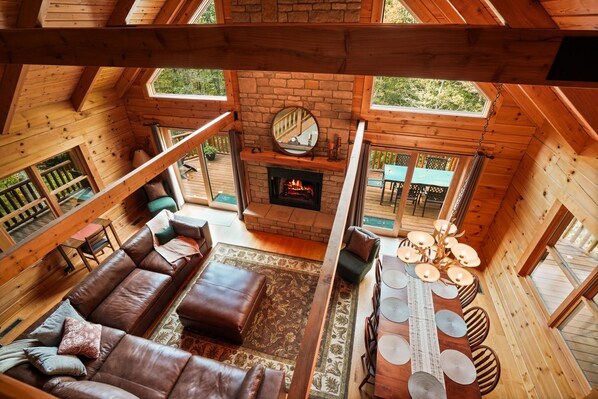 A cabin interior with a fireplace, wooden furniture, and large windows. - Oasis Glen - Views, Pool, Hot Tub (Rockbridge)