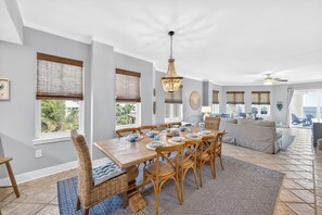 A dining area with a wooden table set for six, surrounded by wicker chairs, a chandelier, and large windows with blinds. - GULF FRONT l BEACH VIEWS FOR DAYS!! l SEASONAL BEACH SETUP INCLUDED! (Santa Rosa Beach)