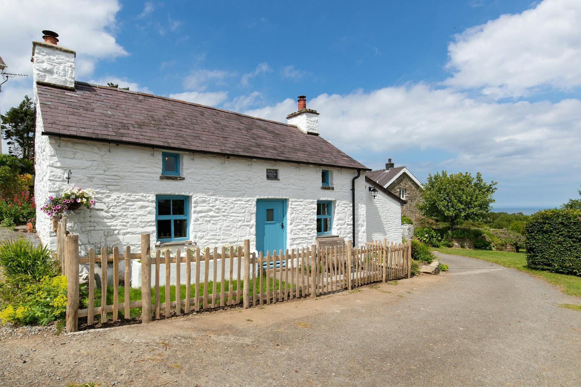 A white stone house with a blue door and windows, a wooden fence, and a gravel driveway.