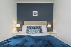 A bed with a blue blanket, two bedside lamps, and a framed picture on the wall. - Tydu View Cottage, New Quay (Llangrannog)