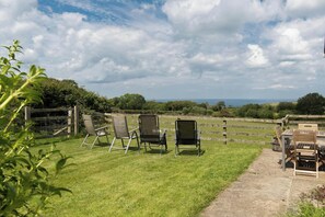 A grassy outdoor area with three chairs and a bench, a wooden fence, and a view of the landscape. - Tydu View Cottage, New Quay (Llangrannog)