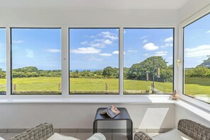 A room with a large window overlooking a green field and trees. - Tydu View Cottage, New Quay (Llangrannog)