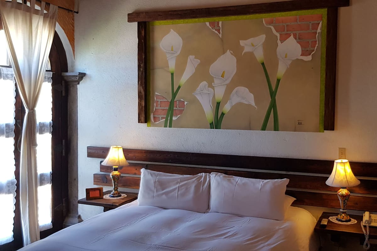A hotel room with a bed, two bedside lamps, a wall-mounted painting of flowers, and a window with curtains.