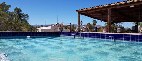 A swimming pool with a tiled edge, a covered area with a roof, and a building in the background.