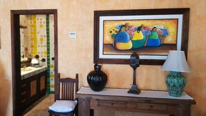 A room with a wooden chair, a small table with a lamp, a framed painting, and a tiled wall with a door. - Posada de las Flores Loreto (Loreto)