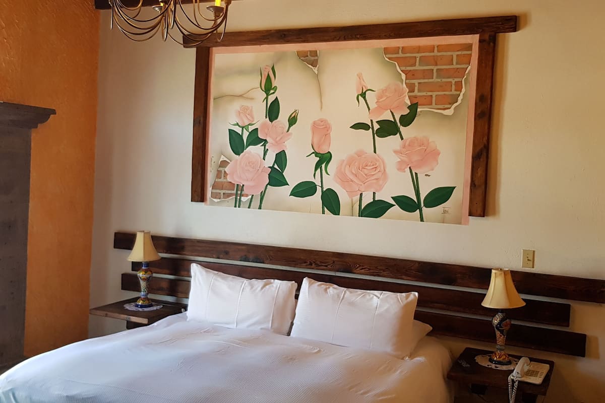 A hotel room with a bed, two bedside tables, a lamp, a painting of roses, and a brick wall.