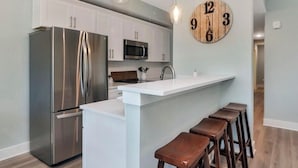 A modern kitchen with stainless steel appliances, a white island with bar stools, and a wooden clock on the wall. - Gulf Front 3 Bedroom Condo in Mexico Beach at The Vue (Mexico Beach)