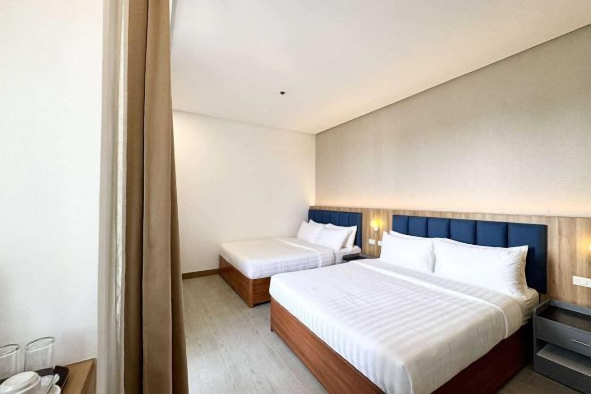 A modern hotel room with a large bed, two smaller beds, a wooden headboard, and a bedside table.