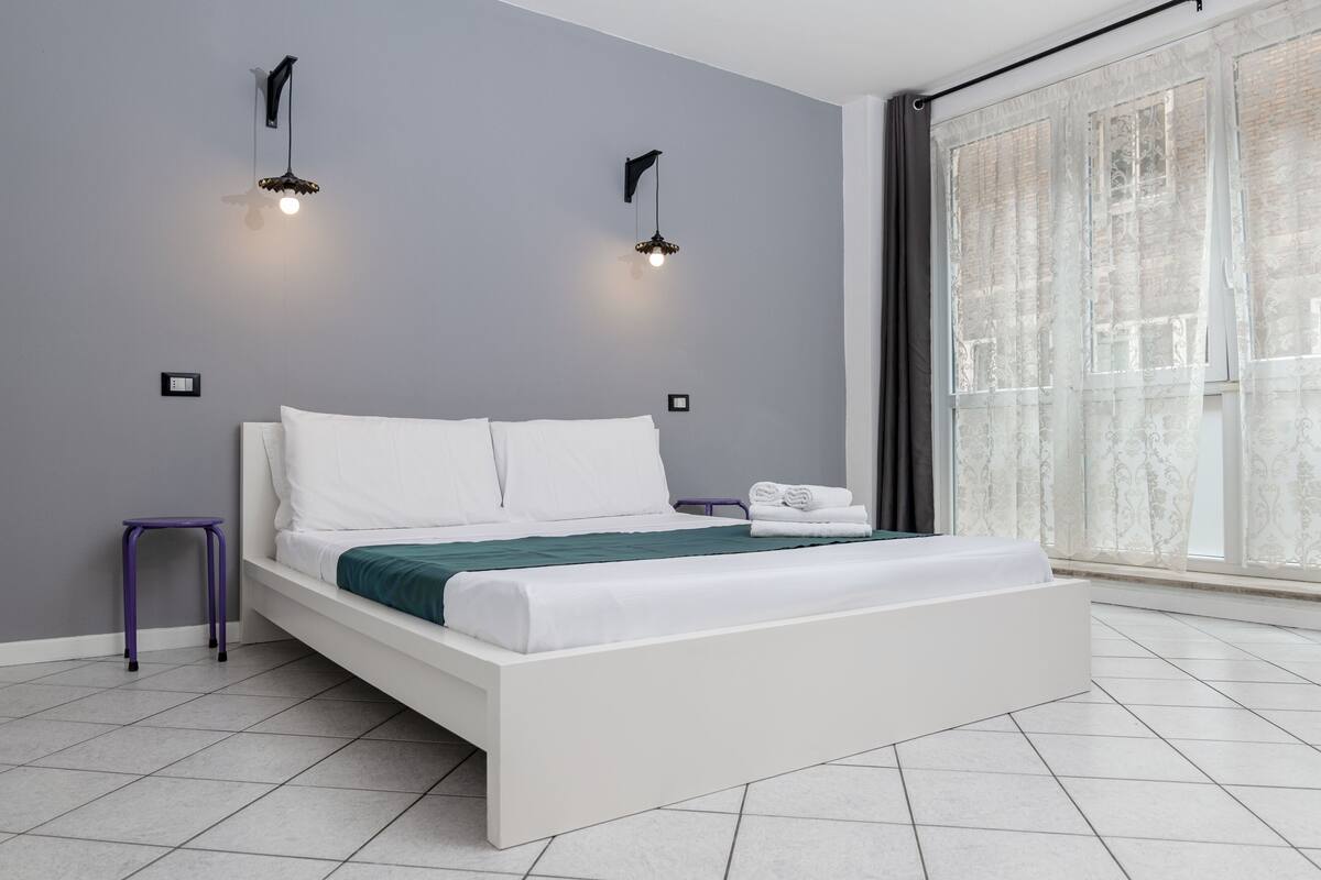 Un dormitori modern amb un llit blanc, parets grises i dues làmpades penjades a la paret.