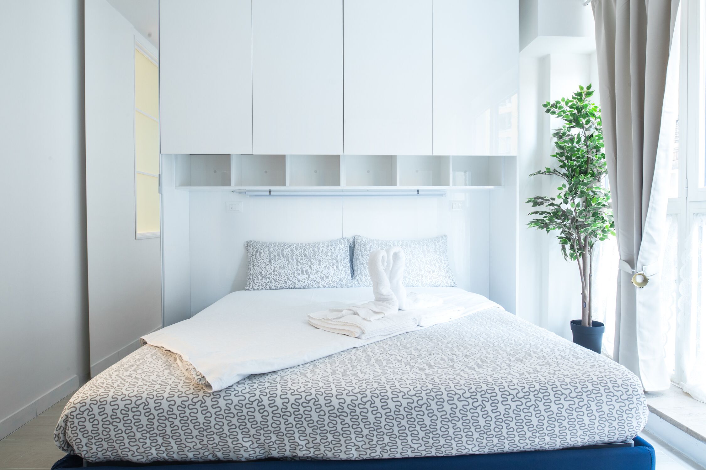 Une chambre à coucher moderne avec un lit, une literie blanche à motifs, une plante en pot et une fenêtre avec des rideaux.