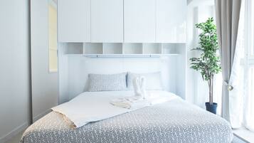 Une chambre à coucher moderne avec un lit, une literie blanche à motifs, une plante en pot et une fenêtre avec des rideaux.