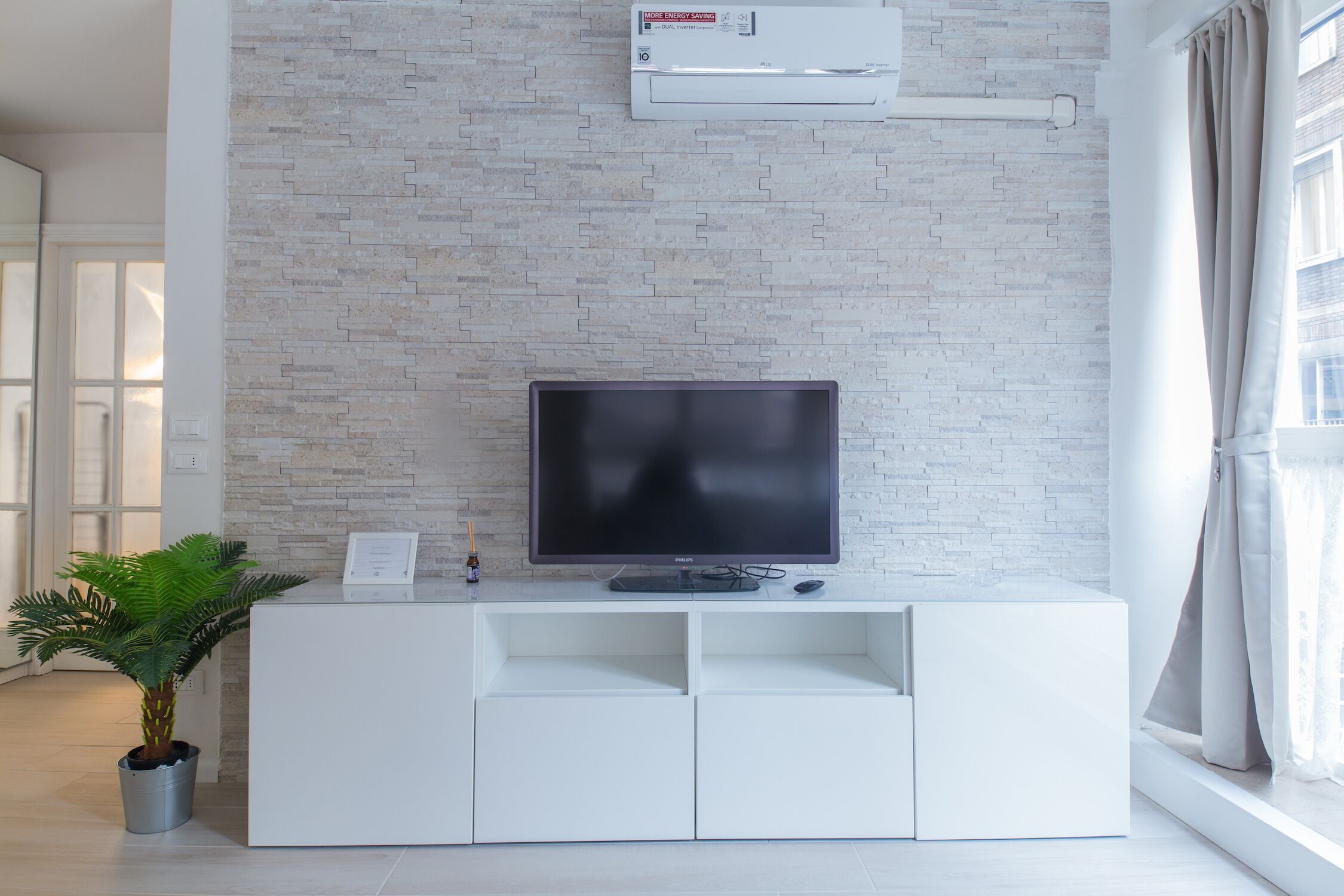 Una sala de estar moderna con un televisor de pantalla plana sobre un mueble blanco, una planta en maceta y una ventana con cortinas.