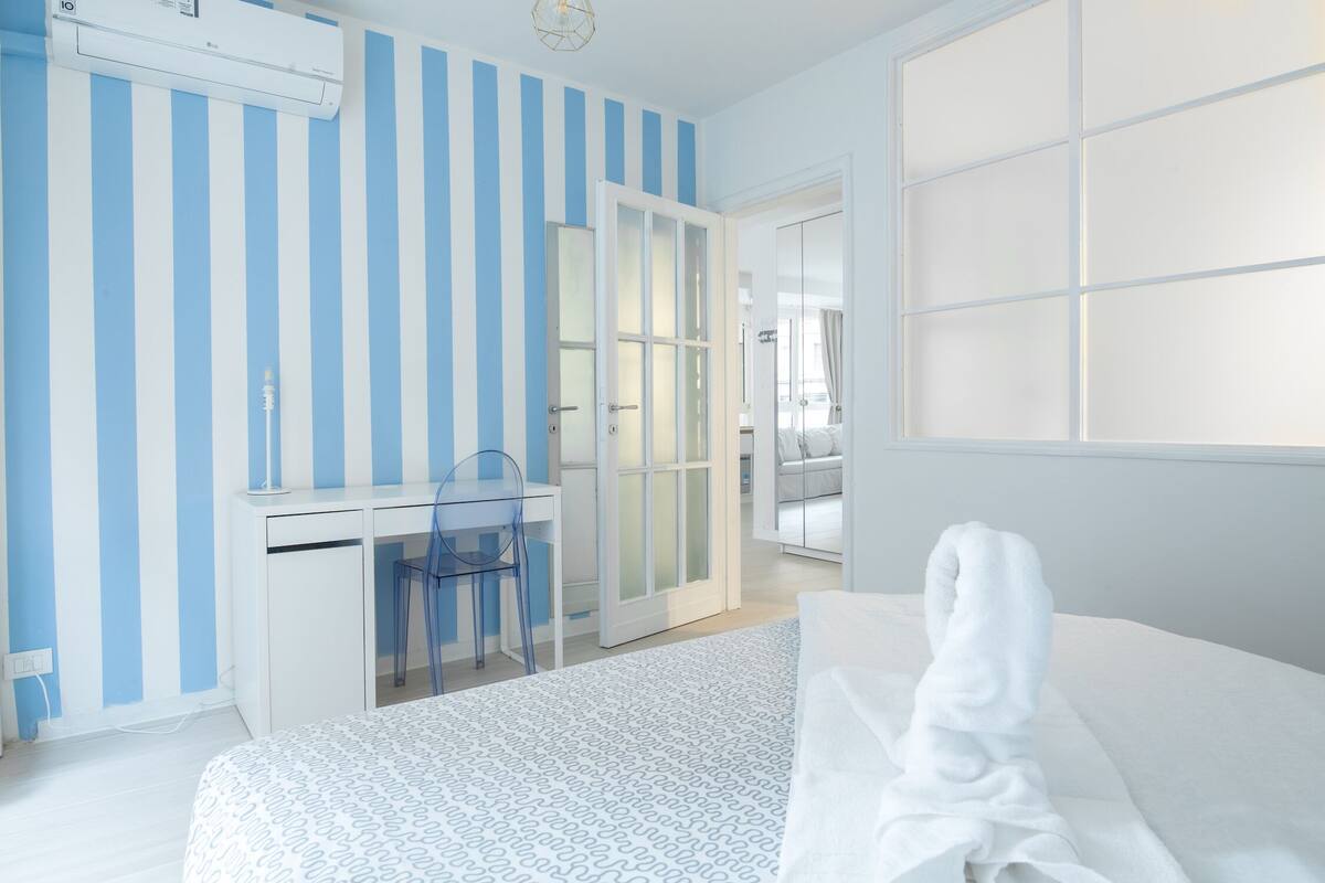 Habitació d'hotel amb un llit, una taula de treball amb una cadira, un armari i una porta corredissa de vidre que dona a un balcó.