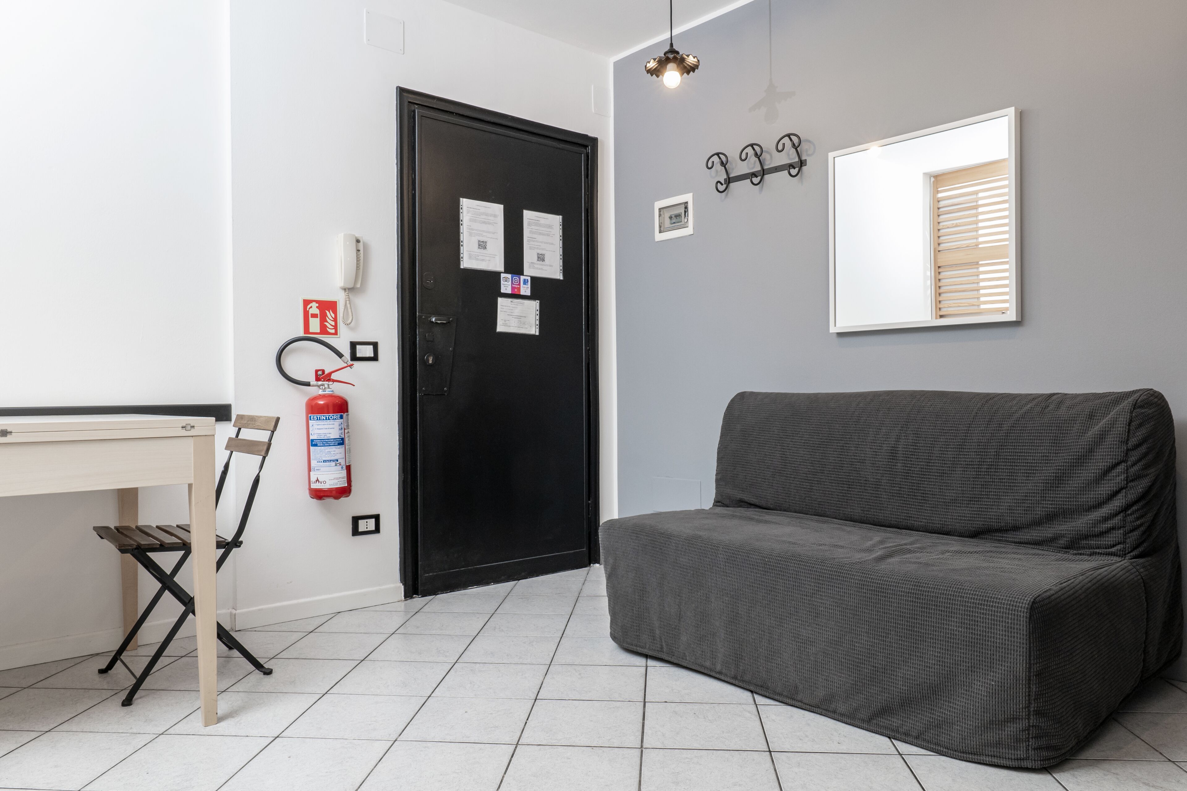 Una habitación con un sofá gris, un escritorio de madera con una silla, una puerta negra con papeles, un extintor y una lámpara fijada a la pared.