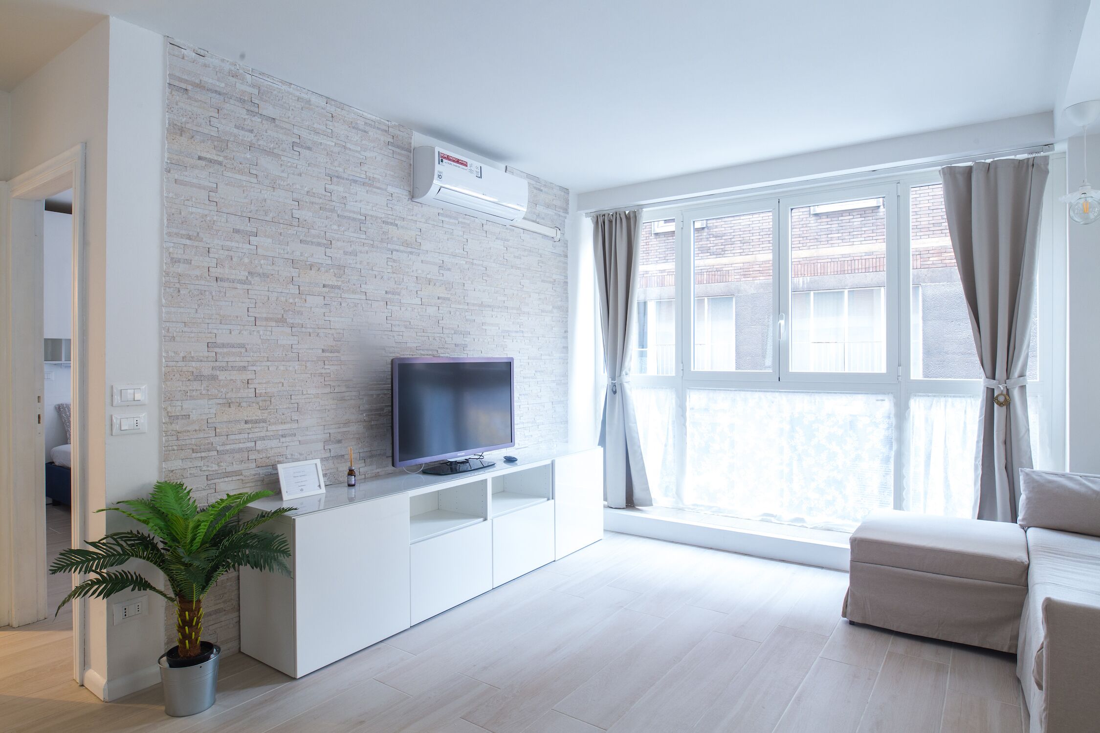 Una sala de estar moderna con un televisor de pantalla plana sobre un mueble blanco, una planta en maceta, un sofá color beige y un ventanal con cortinas.