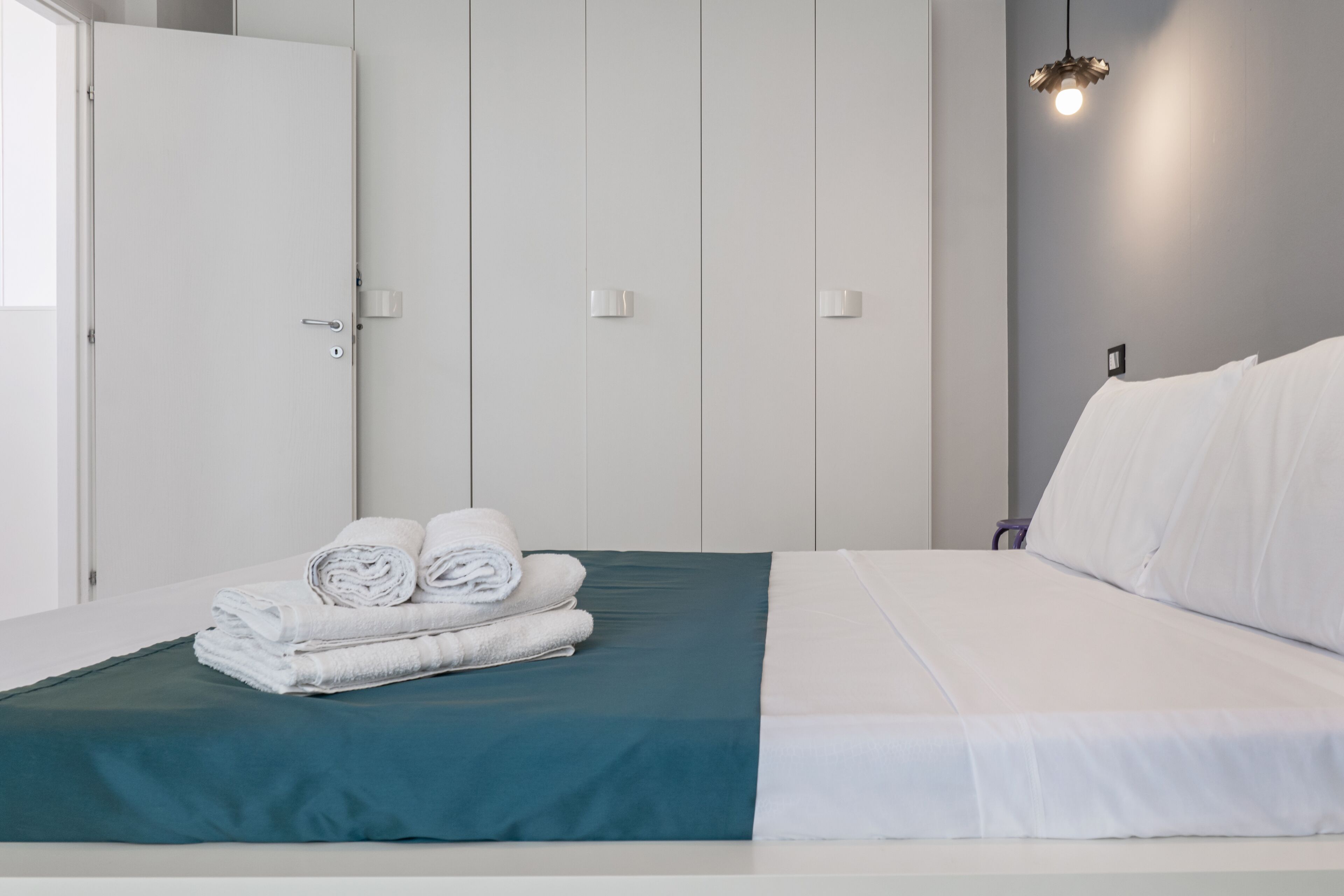 Cama bem arrumada com lençóis brancos e um toque de cor verde-água, um conjunto de toalhas dobradas, uma porta branca e um aplique de luz na parede.