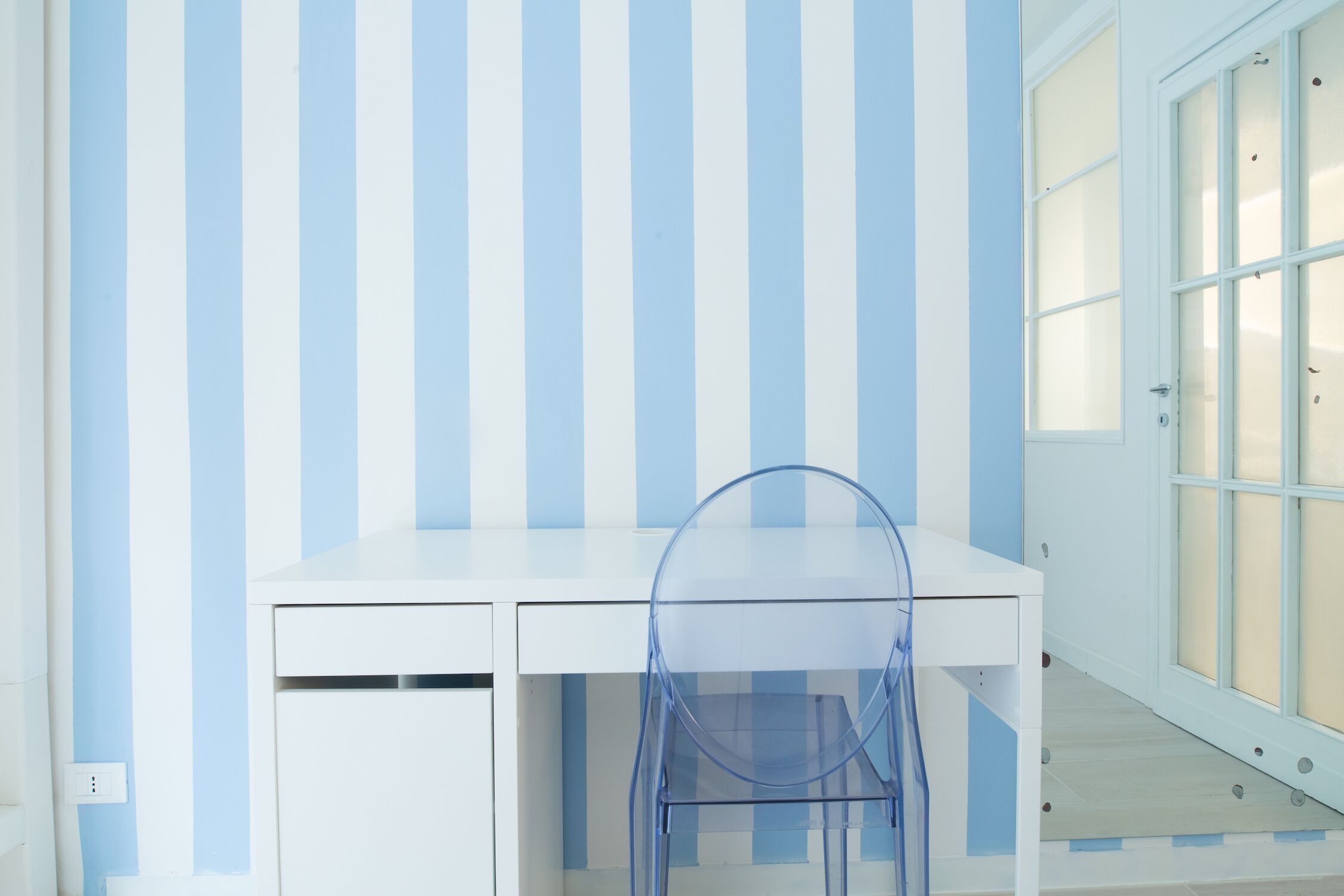 A room with a white desk, a clear plastic chair, and a window with frosted glass.