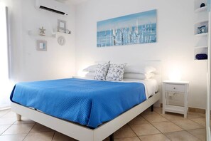 A bedroom with a bed, a nightstand, a wall-mounted air conditioner, and a painting of sailboats.