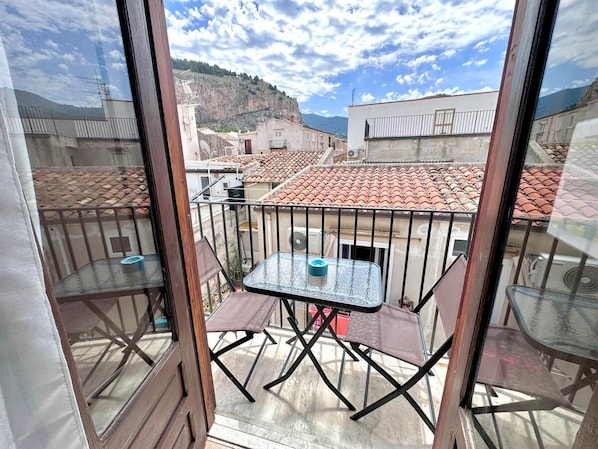 A balcony with a glass table and chairs, overlooking a residential area with red-tiled roofs.